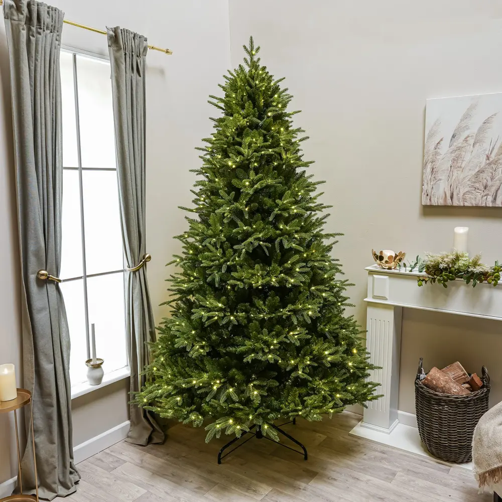 The pre-lit artificial Christmas tree standing in a living room, warm-white lights on, folding metal stand visible
